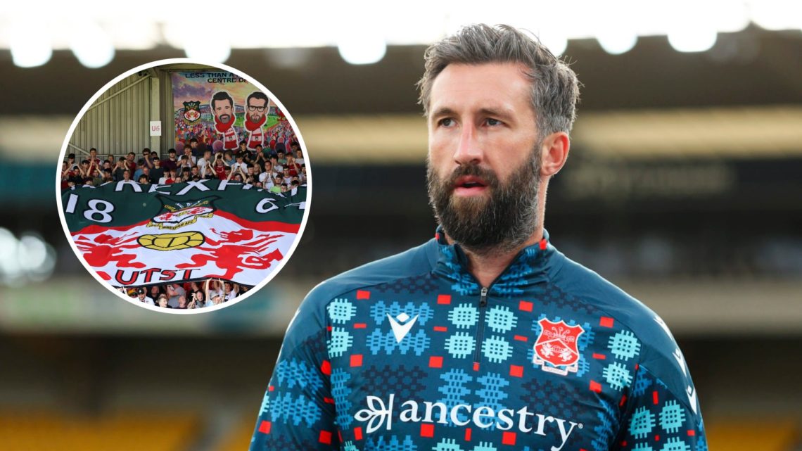 Ollie Palmer of Wrexham AFC warms up. Fans of Wrexham display a flag of their badge with a banner of owners Rob McElhenney and Ryan Reynolds.