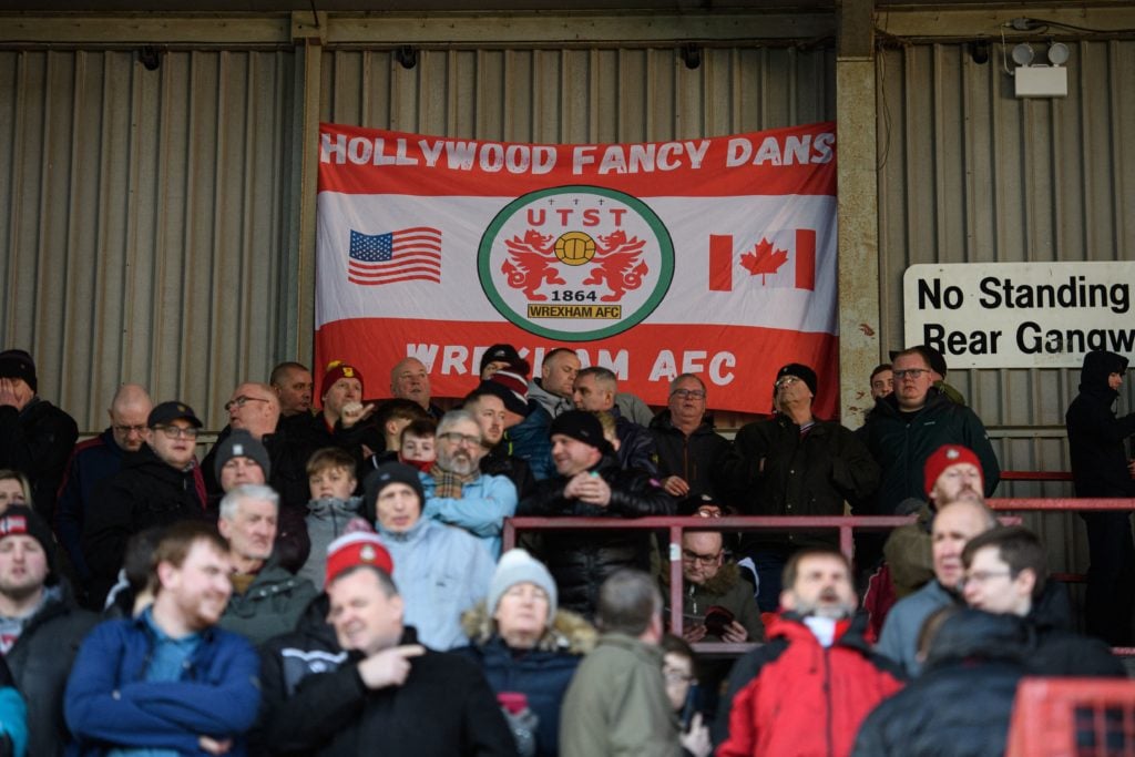Supporters of Wrexham Association Football Club stand in front of a banner making reference to their North American owners